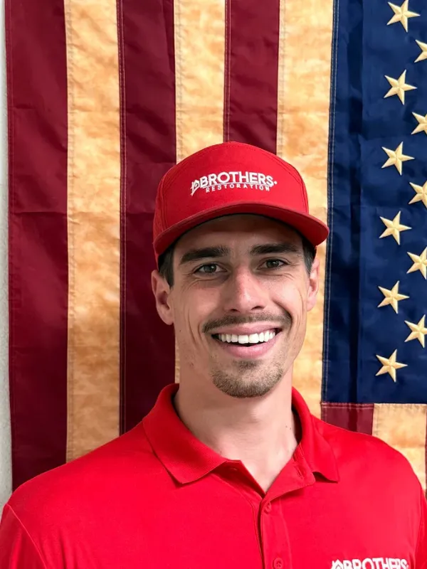 Smiling man in red shirt and cap in front of an American flag.