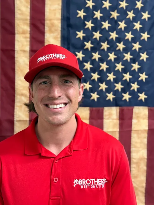 Smiling man in a red Brothers Restoration shirt and cap in front of an American flag.