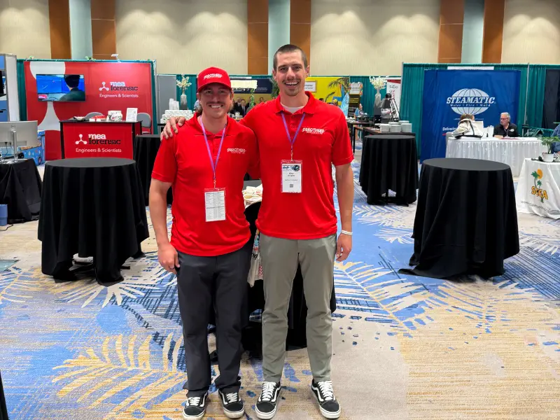Two men in red shirts pose at a trade show booth.