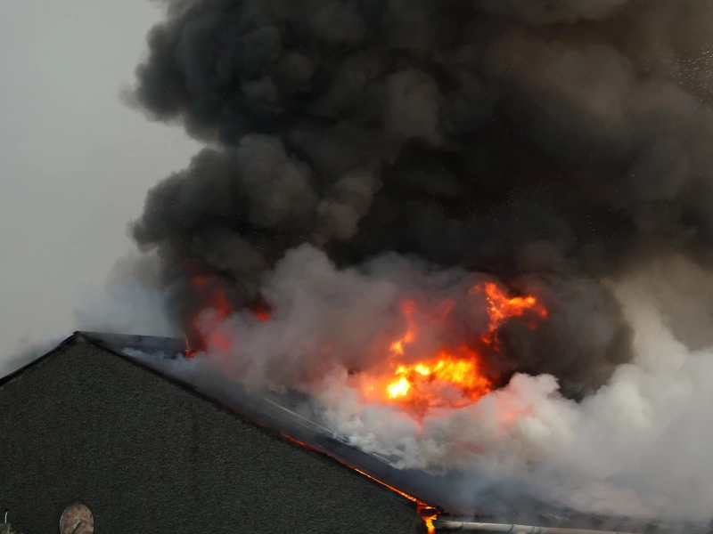Flames and thick black smoke engulf the roof of a building on fire.
