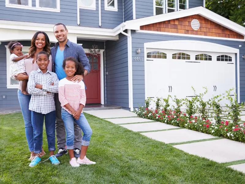 A smiling family of five standing on the lawn in front of a blue house with a white garage door.
