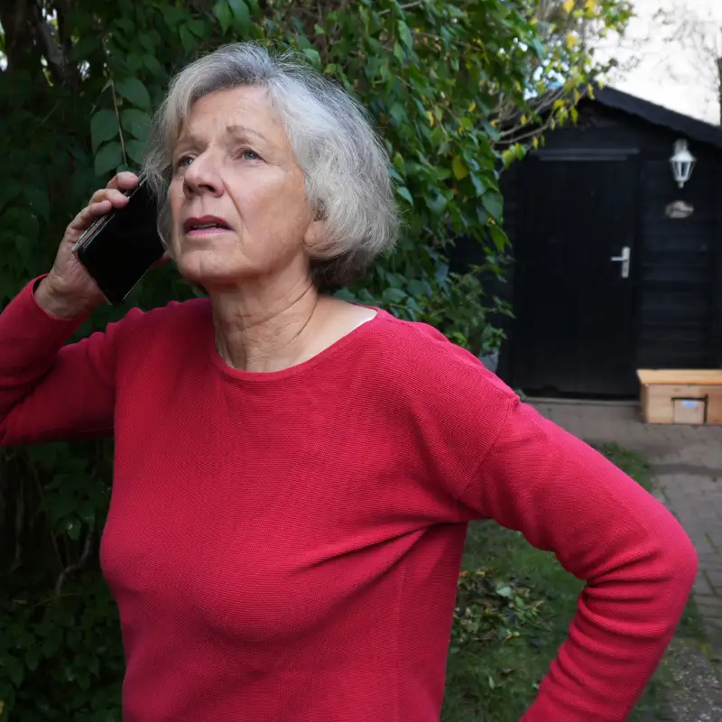 Older woman in a red sweater talking on a phone outdoors near green bushes and a black shed.