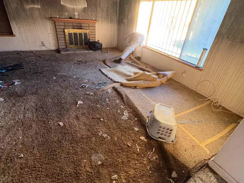 Smoke-damaged living room with peeled carpet and a person near the window.