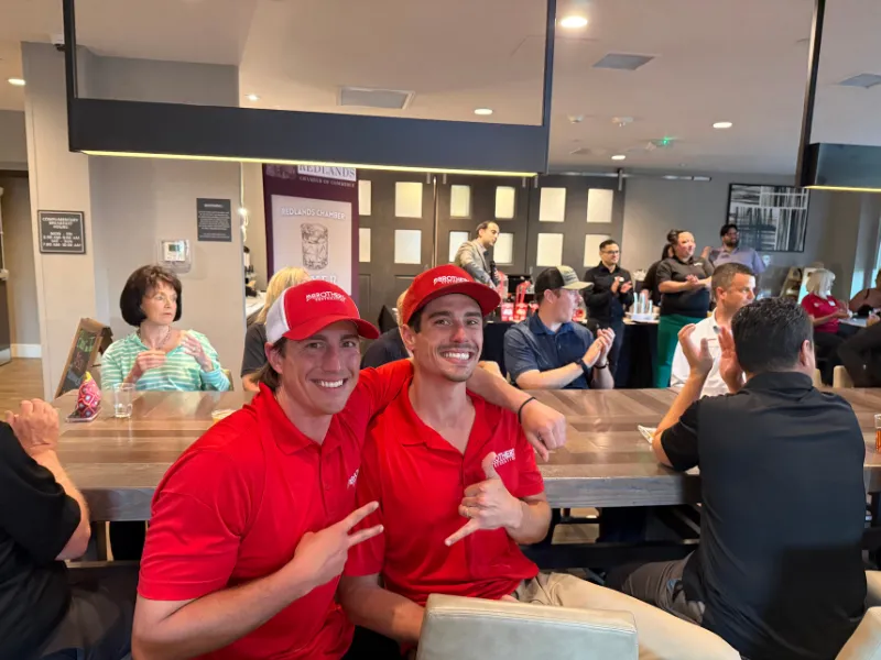 Two men in red shirts and caps pose at a crowded indoor gathering.